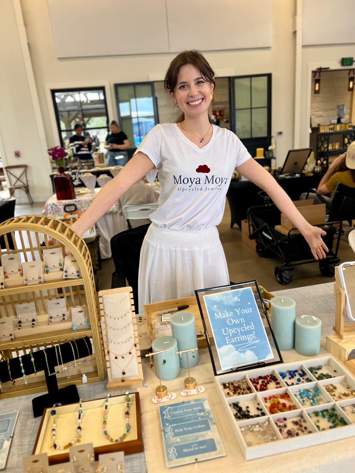Grace at the Moya Moya booth, arms wide behind a table of upcycled jewelry and a 'Make Your Own Upcycled Earrings' sign